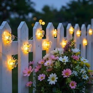 May include: A decorative string of bee-shaped lights adorns a white picket fence. The lights are strung on a thin wire, with a bouquet of pink and white flowers in the foreground. The lights are yellow and the bees have black stripes.