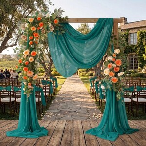 May include: A wooden wedding arch draped with flowing teal fabric and decorated with arrangements of orange and cream-colored roses and green foliage. The arch stands on a wooden platform, framing a stone path leading toward a building.