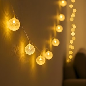 May include: A string of warm white globe lights with a crackle glass effect. The lights are strung on a thin wire, emitting a soft glow. The background is a neutral colour, highlighting the illuminated spheres.