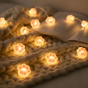 Puede incluir: Una cadena de luces decorativa con bombillas en forma de patas de gato. Cada pata tiene un centro rosa y emite un brillo suave y cálido. Las luces están ensartadas en un cable fino y plateado. Las luces están sobre una manta de punto blanca.