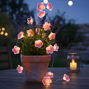 May include: A terracotta pot with lavender and a string of pink flower lights. The lights are wrapped around the plant and create a warm glow. A glass lantern with a lit candle sits nearby on a wooden table.