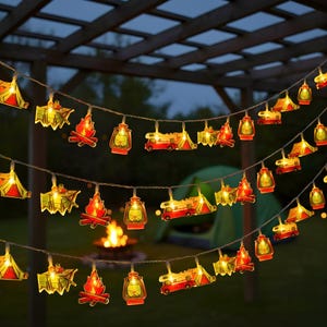 Puede incluir: Luces de cadena decorativas con recortes temáticos de camping, que incluyen tiendas de campaña, fogatas, linternas y autocaravanas. Las luces están ensartadas en un entorno oscuro al aire libre, con una tienda de campaña y una hoguera visibles.