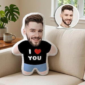 May include: A custom-printed pillow in the shape of a person, featuring a smiling face and the text "I ❤️ YOU" on a black shirt. The pillow has a light blue shorts design and is placed on a beige sofa.