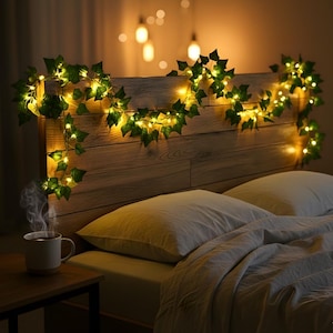 May include: A cozy bedroom scene features a wooden headboard adorned with a garland of green ivy and warm, glowing fairy lights. White pillows and a soft blanket rest on the bed. A steaming mug sits on a small wooden table.