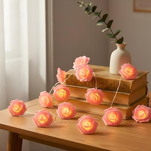 May include: A garland of pink rose fairy lights, each with a warm yellow glow. The lights are on a wooden table, beside a stack of old books and a white vase with greenery.