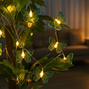 May include: A string of butterfly-shaped fairy lights with a warm glow. The translucent green and blue butterflies are strung on a thin wire, interspersed among green leaves. The lights are illuminated, creating a soft, ambient effect.
