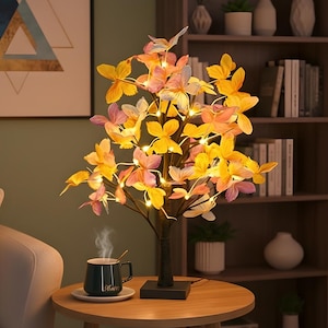May include: A butterfly tree lamp with yellow, pink, and orange fabric butterflies. The lamp has a black base and stem, with small lights illuminating the butterflies. A dark green mug with the word "Home" is on a small table.