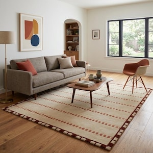 Puede incluir: Una alfombra rectangular con base de color crema que presenta filas horizontales repetidas de peque&ntilde;os guiones rectangulares de color rojo oscuro y un borde a juego, colocada sobre un suelo de madera bajo una mesa de centro.