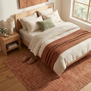 May include: A bedroom featuring a light wood bed frame with beige bedding, a chunky knit rust-coloured throw blanket, and a sage green accent pillow. A rust-coloured area rug with white dotted patterns lies beneath the bed, matching the wall hanging.