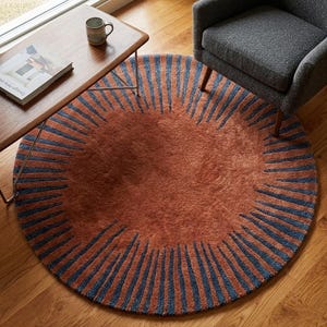 May include: A round area rug featuring a solid terracotta-colored center surrounded by a border of radiating dark blue stripes. The rug has a textured, tufted pile surface and is positioned on a wooden floor, partially underneath a wooden table.