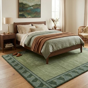 May include: A bedroom with a wooden bed, sage green bedding, and a rust-coloured throw. A green rug with a geometric border is on the wooden floor. A landscape painting hangs above the bed, creating a calming atmosphere.