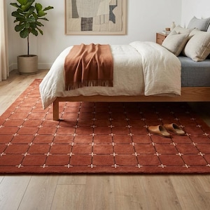 Puede incluir: Una alfombra de color terracota con un patrón de cuadrícula de cruces blancas en un dormitorio. La alfombra está colocada debajo de una cama con estructura de madera, un edredón blanco y una manta marrón. La habitación tiene pisos de madera y una planta en maceta.