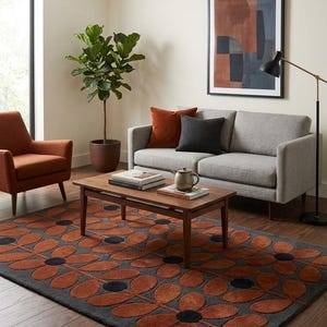 May include: A rectangular area rug featuring a repeating pattern of dark gray flower shapes with burnt orange petals. The rug is positioned on a dark wood floor in a room containing a gray sofa, an orange armchair, a wooden coffee table, and a plant.