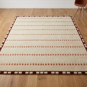 Puede incluir: Una alfombra rectangular con una base de color crema y filas horizontales de peque&ntilde;os detalles rectangulares en color terracota. La alfombra est&aacute; enmarcada por un borde marr&oacute;n oscuro con un patr&oacute;n repetitivo de peque&ntilde;os cuadrados claros.