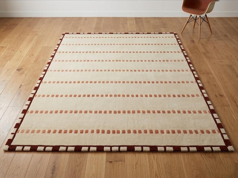 Puede incluir: Una alfombra rectangular con una base de color crema y filas horizontales de peque&ntilde;os detalles rectangulares en color terracota. La alfombra est&aacute; enmarcada por un borde marr&oacute;n oscuro con un patr&oacute;n repetitivo de peque&ntilde;os cuadrados claros.
