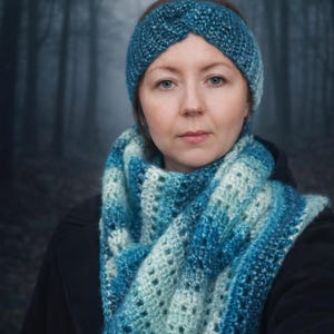 Op de afbeelding: Een vrouw draagt een turquoise gehaakte hoofdband en een bijpassende sjaal. De sjaal heeft een gradiëntpatroon van licht- en donkerblauw. De achtergrond is een vage bosscène.
