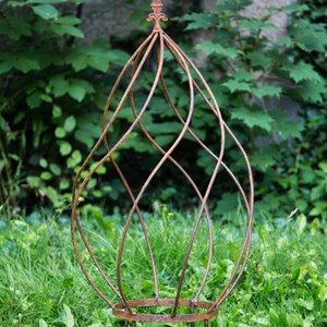 Könnte beinhalten: Dekorative Gartenobelisk aus rostbraunem Metall. Der Obelisk hat ein käfigartiges Design mit geschwungenen Linien und einem Fleur-de-Lis-Aufsatz. Er steht auf kurzen Beinen in einem grasbewachsenen Bereich.