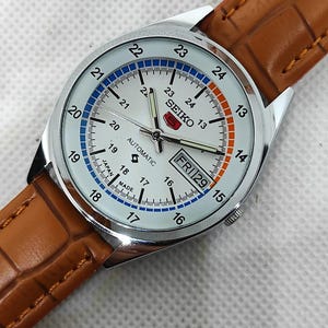 May include: Silver-toned Seiko automatic watch with a white face, featuring blue and orange accents. The watch has a brown leather band. The face displays the day and date, with the text "FRI 29".