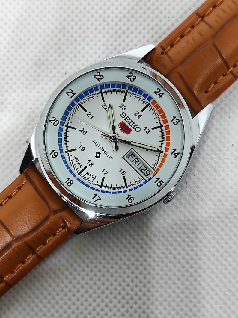 May include: Silver-toned Seiko automatic watch with a white face, featuring blue and orange accents. The watch has a brown leather band. The face displays the day and date, with the text "FRI 29".