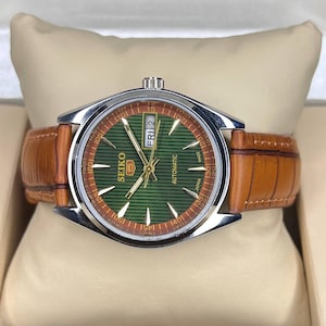 May include: A Seiko 5 automatic watch with a green dial and brown leather strap. The watch face features gold hands and markers, with the day and date displayed. The watch has a silver-colored case and a brown leather band.