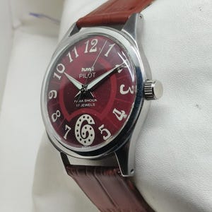 May include: A wristwatch with a silver-tone case and a textured brown leather strap. The dial is deep red with white Arabic numerals, silver hands, and the text HMT PILOT, PARA SHOCK, 17 JEWELS, and INDIA printed in white on the face.