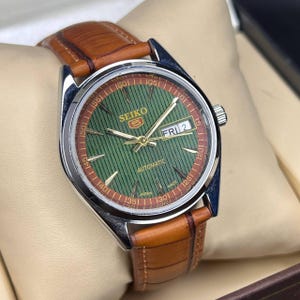 May include: A Seiko 5 automatic watch with a green dial and brown leather strap. The watch face displays the day and date, with gold-colored hands and markers. The watch has a stainless steel case and a brown leather band.