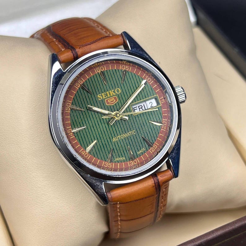 May include: A Seiko 5 automatic watch with a green dial and brown leather strap. The watch face displays the day and date, with gold-colored hands and markers. The watch has a stainless steel case and a brown leather band.