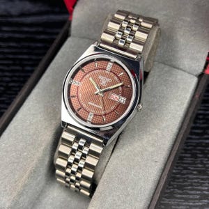 Op de afbeelding: Een zilverkleurig Seiko 5 automatisch polshorloge met een getextureerde koperkleurige wijzerplaat, zilveren wijzers en uurmarkeringen. Het horloge heeft een dag-datumvenster op de drie-uurpositie en een metalen schakelarmband.