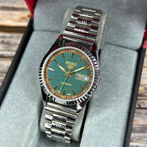 May include: A Seiko 5 automatic wristwatch with a silver stainless steel band. The watch face is green with gold hands and markers, and the day and date are displayed. The watch is presented in a black box with a gray interior.