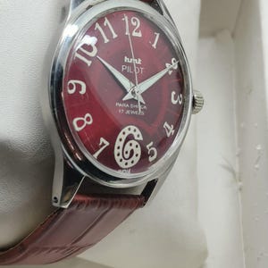 May include: A wristwatch with a round silver-tone case and a deep red dial featuring white Arabic numerals, including a stylized number six. The dial displays the text HMT PILOT, PARA SHOCK, 17 JEWELS, and INDIA. It has a brown leather strap.