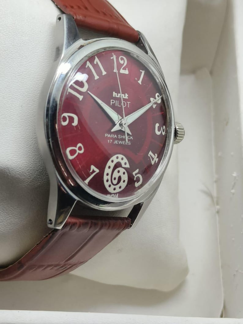 May include: A wristwatch with a round silver-tone case and a deep red dial featuring white Arabic numerals, including a stylized number six. The dial displays the text HMT PILOT, PARA SHOCK, 17 JEWELS, and INDIA. It has a brown leather strap.