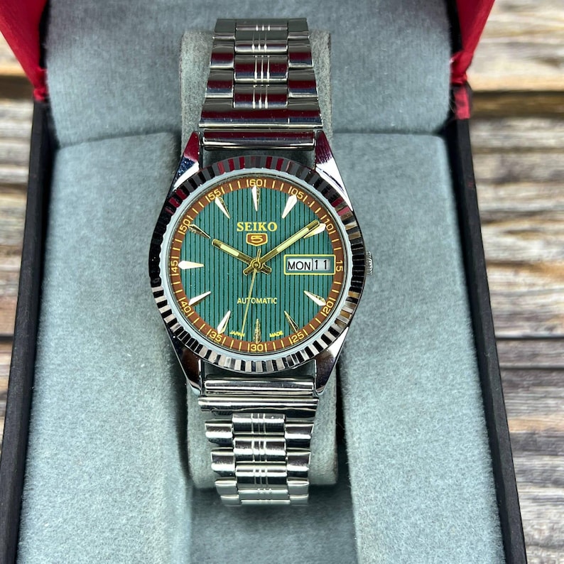 May include: A silver-toned Seiko 5 automatic watch with a green dial and a day-date display. The watch features a fluted bezel and a stainless steel bracelet. The dial has gold-toned hands and markers. The watch is presented in a black and red box.