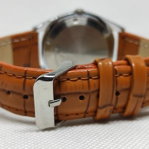 May include: Close-up of a wristwatch with a brown leather band. The band has a textured, crocodile-like pattern and a silver buckle. The watch face is visible in the background.