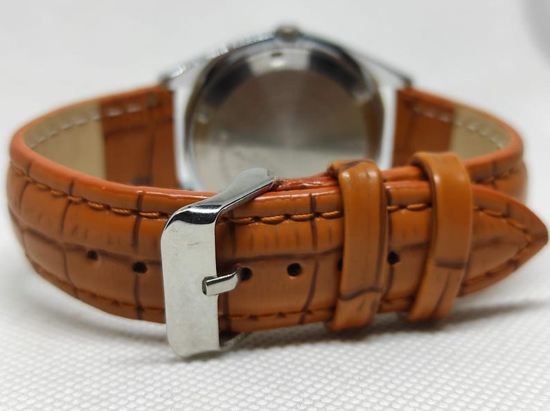 May include: Close-up of a wristwatch with a brown leather band. The band has a textured, crocodile-like pattern and a silver buckle. The watch face is visible in the background.
