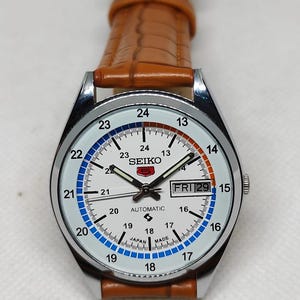 May include: A silver-toned Seiko automatic watch with a white face, featuring black hands and hour markers. The watch has a brown leather strap and displays the day and date. The face has blue and orange accents.