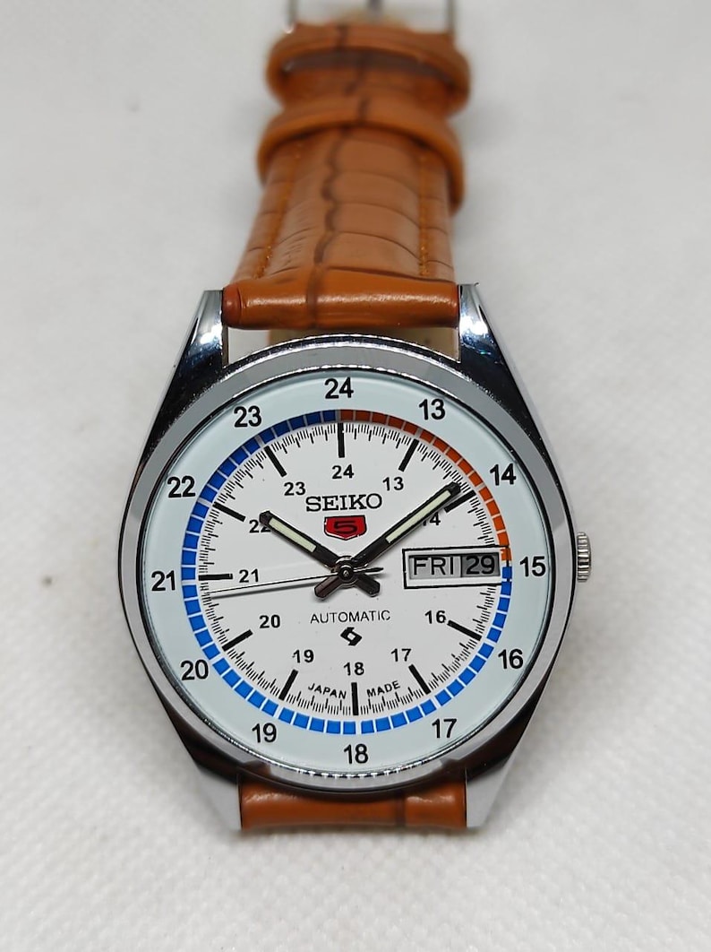 May include: A silver-toned Seiko automatic watch with a white face, featuring black hands and hour markers. The watch has a brown leather strap and displays the day and date. The face has blue and orange accents.