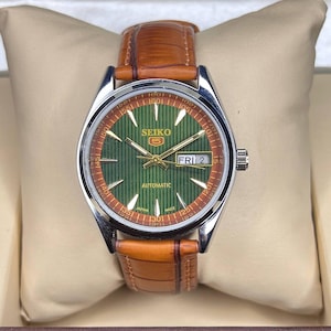 May include: A Seiko 5 automatic watch with a green dial and brown leather strap. The watch face displays the day and date, with gold-tone hands and markers. The watch is presented in a beige box.