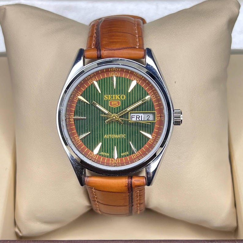 May include: A Seiko 5 automatic watch with a green dial and brown leather strap. The watch face displays the day and date, with gold-tone hands and markers. The watch is presented in a beige box.