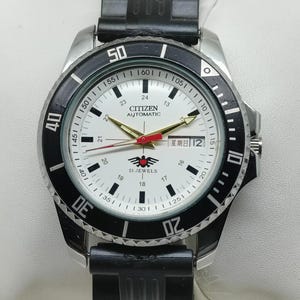 May include: A Citizen Automatic watch with a white face, black bezel, and black rubber strap. The watch features gold hands, a red second hand, and the text "CITIZEN AUTOMATIC" and "21 JEWELS". The day of the week is displayed in Japanese.