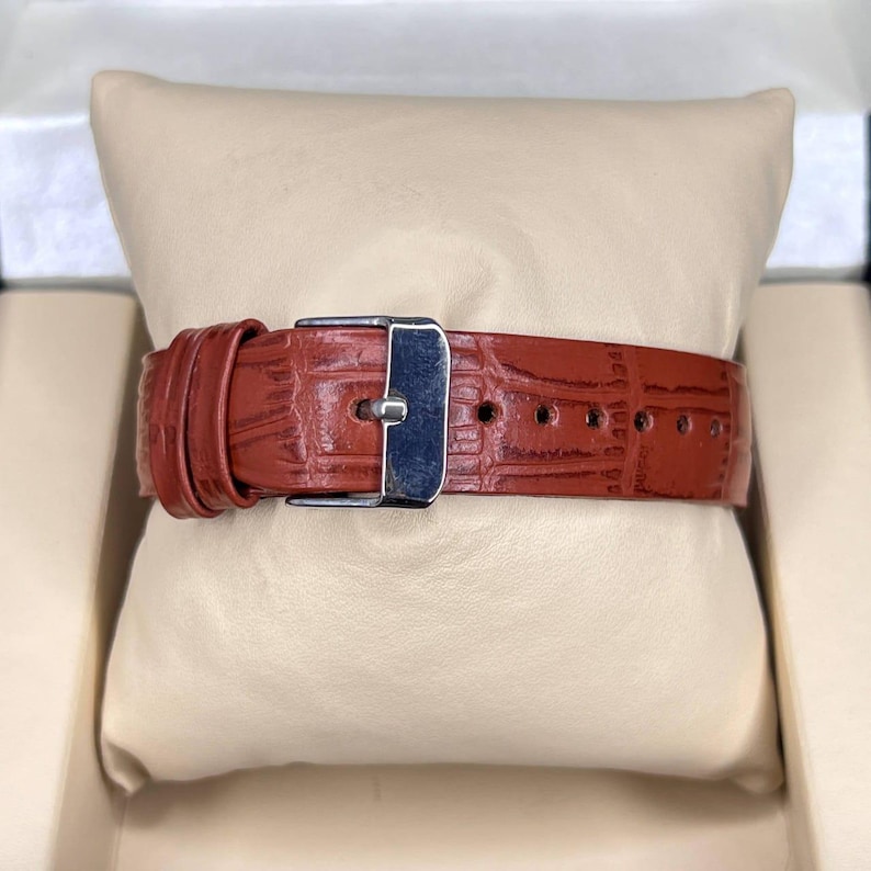 May include: Close-up of a watch band made of reddish-brown leather with a crocodile skin texture. The band has a silver-colored buckle and several holes for adjustment. The watch band is resting on a beige pillow.