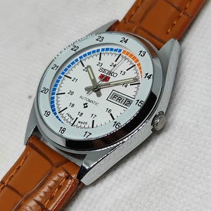 May include: A silver-toned Seiko automatic watch with a white face, day/date display, and a brown leather strap. The watch face features blue and orange accents, with the day displayed as FRI 2. The watch is marked "JAPAN MADE".