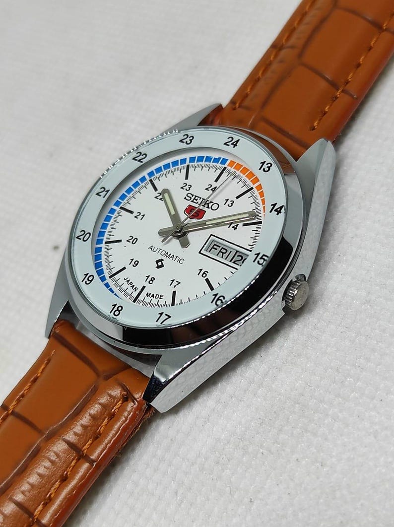 May include: A silver-toned Seiko automatic watch with a white face, day/date display, and a brown leather strap. The watch face features blue and orange accents, with the day displayed as FRI 2. The watch is marked "JAPAN MADE".