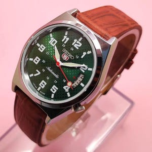Puede incluir: Un reloj automático Seiko con esfera verde y correa de cuero marrón. El reloj tiene números blancos, una pantalla de fecha y un segundero rojo. La caja del reloj es plateada.