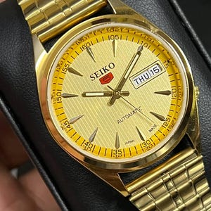 May include: A gold-toned Seiko 5 automatic wristwatch. The watch features a gold dial with a day-date display, gold hands, and a gold-colored bracelet. The watch face has the word "SEIKO" and "AUTOMATIC" printed on it.