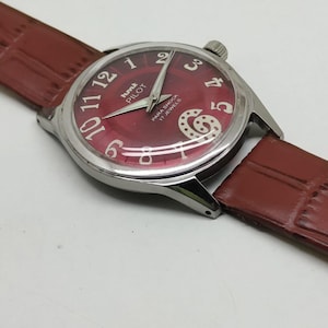 May include: A wristwatch with a round silver-tone case and a deep red dial featuring white Arabic numerals. The dial displays the text HMT PILOT and PARA SHOCK 17 JEWELS. It includes silver-tone hands and a brown textured leather strap.