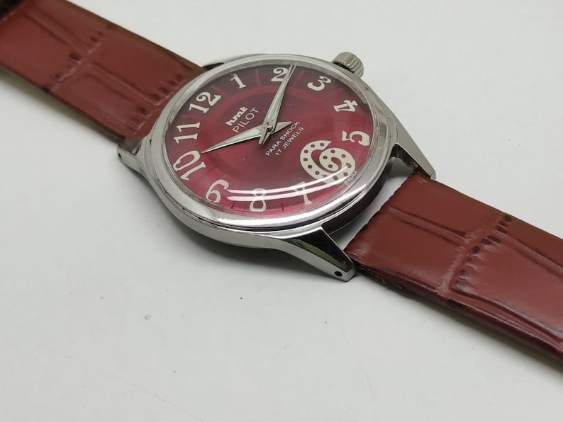 May include: A wristwatch with a round silver-tone case and a deep red dial featuring white Arabic numerals. The dial displays the text HMT PILOT and PARA SHOCK 17 JEWELS. It includes silver-tone hands and a brown textured leather strap.