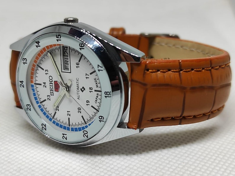 May include: A silver-toned Seiko automatic watch with a white face, featuring black numerals and a day-date display. The watch has a brown leather band with a crocodile pattern. The watch face has blue and orange accents.