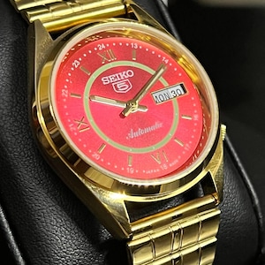 May include: A gold-coloured Seiko 5 automatic wristwatch with a red dial. The watch face features Roman numerals, a day-date display, and the Seiko logo. The watch band is also gold-coloured.