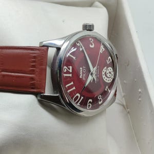 May include: A wristwatch with a circular silver-tone case and a deep red dial featuring white Arabic numerals. The watch face displays the text HMT PILOT and 17 JEWELS. It is fitted with a brown leather strap with a textured crocodile-like pattern.
