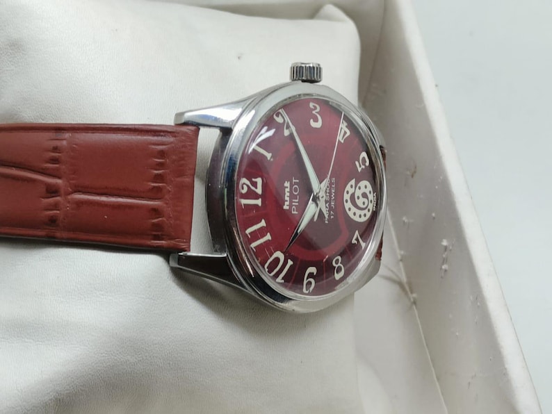May include: A wristwatch with a circular silver-tone case and a deep red dial featuring white Arabic numerals. The watch face displays the text HMT PILOT and 17 JEWELS. It is fitted with a brown leather strap with a textured crocodile-like pattern.
